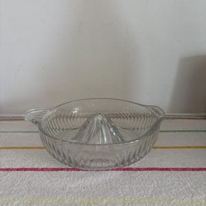 May include: A clear glass citrus juicer with a ribbed bowl and a central reamer. The juicer has a handle on each side. The design is simple and functional, ideal for squeezing fresh juice. A kitchen tool for preparing beverages.