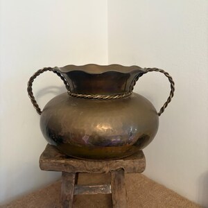 May include: A brass vase with a hammered finish, round body, and a ruffled rim. It features two twisted rope handles. The vase is displayed on a small, rustic wooden stool. The vase has a warm, metallic finish.