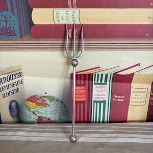 Puede incluir: Un tenedor de plata de tres puntas con un mango redondo. El tenedor está apoyado sobre una superficie de tela a rayas. El tenedor está delante de una pila de libros con texto en francés en los lomos.