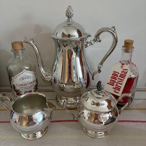 May include: A silver tea set comprising a teapot, creamer, and sugar bowl. Two bottles are also present, one with a cork stopper and the other with a "happy christmas" tag. The set is displayed on a striped tablecloth.