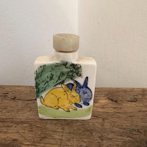 Puede incluir: Una pequeña botella de cerámica rectangular con un tapón de color claro. La botella presenta una escena pintada a mano de dos conejos, uno amarillo y otro azul, bajo un árbol verde. La botella está sobre una superficie de madera.