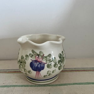May include: A small, white ceramic creamer with a scalloped rim. It features a floral design with green leaves and a purple and pink flower.