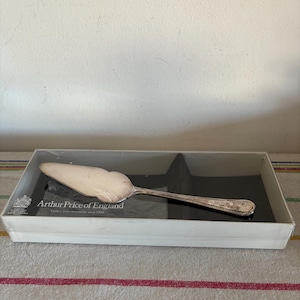 May include: A silver cake server with a scalloped edge, presented in a white box. The box features the text "Arthur Price of England." The server has a decorative handle and sits on a black surface.