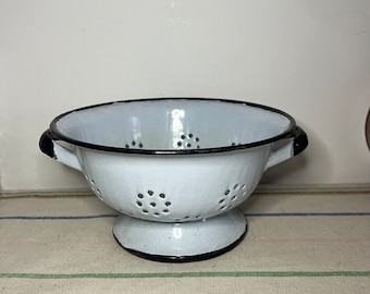 May include: White enamel colander with black rim and handles. The colander has a footed base and a pattern of small holes.