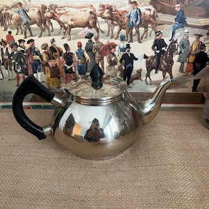 Puede incluir: Una tetera de plata con un asa negra. La tetera tiene un cuerpo redondo y un pico en el lateral. La tetera está sobre una superficie de arpillera marrón.