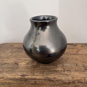 Peut inclure: Un petit vase en céramique arrondi avec une finition métallique gris acier. Le vase présente une surface lisse et réfléchissante avec un corps large et bulbeux qui se rétrécit vers une ouverture circulaire étroite au sommet, avec un vernis brillant.