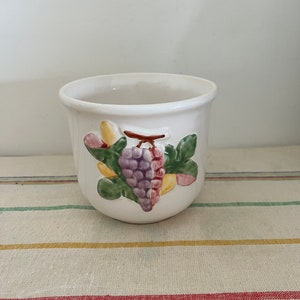 Puede incluir: Maceta de cerámica blanca con un diseño de uvas. Las uvas son moradas, verdes y amarillas. La maceta tiene una forma redondeada.