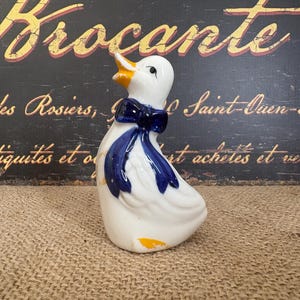 May include: A white ceramic duck figurine featuring a glossy finish, a bright yellow beak, and a dark blue bow tied around its neck. The duck is depicted in a standing position with its head tilted upward, showcasing smooth, rounded contours.