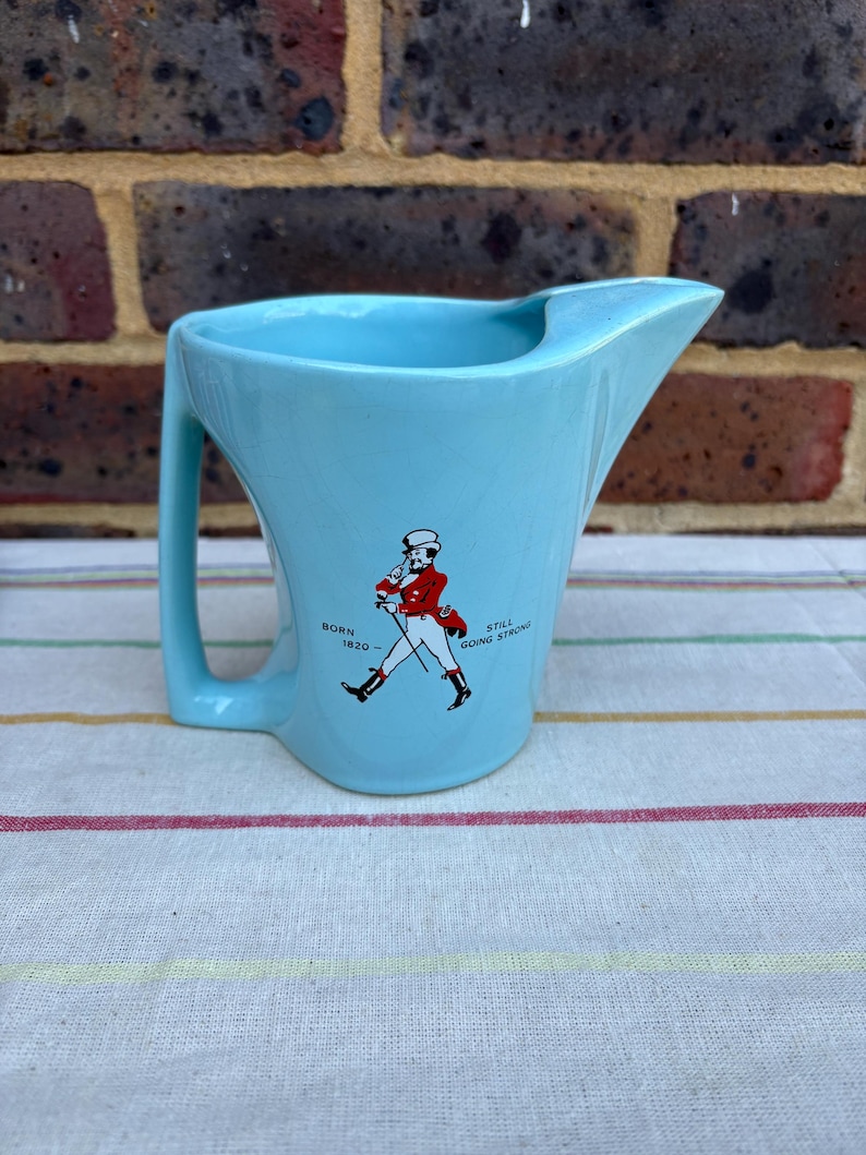 May include: A light blue ceramic jug with a handle and a spout. It features a red and white illustration of a man walking with a cane and the text "Born 1820 - Still Going Strong".