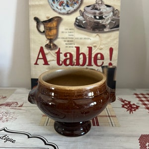 Op de afbeelding: Een bruine keramische kom met twee handvatten, op een voetstuk. De kom staat voor een boek met de titel "A table!" met afbeeldingen van antiek servies. De kom heeft een geglazuurde afwerking.