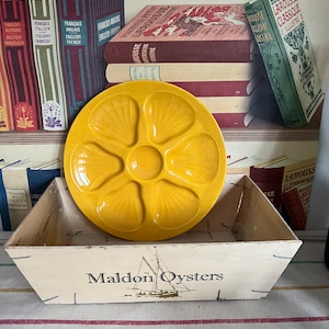 Puede incluir: Un plato de ostras de cerámica amarillo con seis hendiduras individuales se encuentra sobre una caja de madera con las palabras "Maldon Oysters" impresas en la parte delantera. La caja está rodeada de una variedad de libros antiguos con títulos en francés e inglés.
