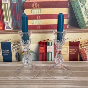 Op de afbeelding: Twee heldere glazen kaarsenstandaards met een diamantpatroon op de basis en een ronde bovenkant. Elke standaard heeft een donkerblauwe kaars erin. De kaarsenstandaards staan op een gestreept tafelkleed voor een boekenkast met boeken in het Frans en Engels.