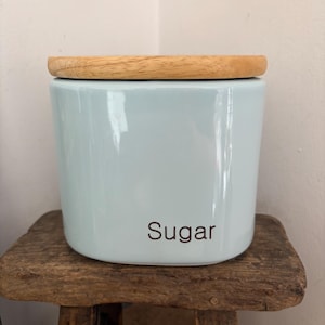 May include: A light blue ceramic canister with a smooth, glossy finish and a natural wood lid. The word "Sugar" is printed in a simple, dark brown sans-serif font on the front of the canister, which has a cylindrical shape with rounded edges.