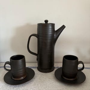 Puede incluir: Un juego de café de cerámica marrón oscuro con una cafetera alta con asa y boquilla, y dos tazas a juego con platillos. El juego se coloca sobre una superficie a rayas blancas y azules, creando una estética rústica y elegante.