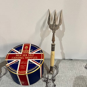 Könnte beinhalten: Eine runde Dose mit Milch-Meersalz-Karamelltrüffeln mit einem Union-Jack-Design. Die Dose ist 8,4 Unzen schwer und wird von Charbonnel et Walker hergestellt, gegründet 1875. Neben der Dose liegt eine silberne Gabel mit einem Knochengriff.