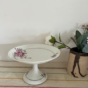 May include: White ceramic pedestal cake stand with a floral design featuring pink roses. The stand has a silver trim around the edge of the plate and the base.