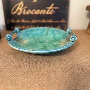 Könnte beinhalten: Eine türkisfarbene und grüne Keramikservierschale mit zwei Griffen. Die Schale hat ein gesprenkeltes Muster und einen leicht erhöhten Rand. Der Hintergrund enthält ein Schild mit den Worten "Antiquités Brocante" in goldfarbenen Buchstaben.