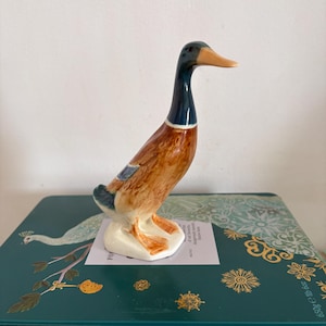 Könnte beinhalten: Eine Keramik-Entenfigur mit braunem Körper, blauem und schwarzem Hals und orangefarbenem Schnabel. Die Ente steht auf einem weißen Sockel. Die Figur steht auf einer tealfarbenen dekorativen Box mit goldenen und weißen floralen Designs.