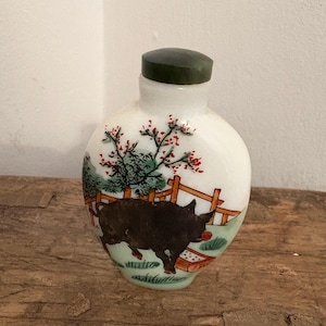 Puede incluir: Una pequeña botella redonda blanca con un tapón verde oscuro. La botella presenta una escena pintada a mano de un cerdo negro en un campo con una valla de madera y árboles. La botella está sobre una superficie de madera.