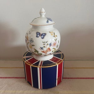May include: A white porcelain jar with a lid, decorated with floral and butterfly patterns. The jar is placed on a box with a Union Jack design in red, white, and blue. The jar has a gold trim.