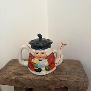 Könnte beinhalten: Eine Keramik-Teekanne in Form eines Mannes mit schwarzem Hut und rotem Mantel. Die Teekanne hat einen weißen Griff und Ausguss mit orangefarbenen Verzierungen. Das Gesicht des Mannes ist auf die Vorderseite gemalt. Die Teekanne steht auf einem Holzschemel.