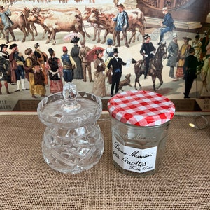 May include: Two glass jars on a hessian surface. One is a clear, cut-glass jar with a lid. The other is a small jar with a red and white gingham lid and a label reading "Bonne Maman des Griottes Fruits Choisis". A vintage print is in the background.