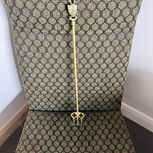 Könnte beinhalten: Ein goldfarbener Metall-Kaminhaken mit einem dekorativen Schild oben und einer dreizackigen Gabel unten. Der Kaminhaken liegt auf einem gemusterten Stuhl mit einem schwarz-goldenen Design.