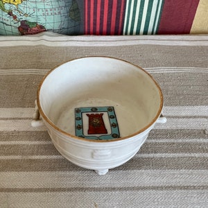 May include: White ceramic bowl with a gold rim and three feet. The bowl has a small, colourful image of a castle with a lion inside on the bottom.