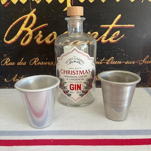 May include: A clear glass gin bottle with a cork stopper and a label that reads "CHRISTMAS CINNAMON GINGER & CARDAMOM GIN". Two small silver metal cups sit in front of the bottle. The background is a vintage sign.