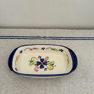 Puede incluir: Un pequeño plato rectangular de cerámica con base blanca y borde azul. El plato está decorado con un diseño floral en azul, rosa, amarillo y verde. El plato mide aproximadamente 15 cm de largo y 7,5 cm de ancho.