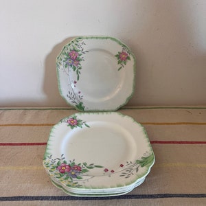Pode incluir: Um conjunto de quatro pratos quadrados de cerâmica branca com um design floral verde. Os pratos têm uma borda festonada e apresentam flores rosa, roxas e amarelas com folhas verdes.