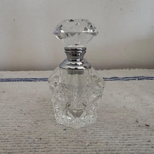 Könnte beinhalten: Ein klarer, facettierter Glasparfümflakon mit einem diamantförmigen Verschluss. Die Flasche hat einen silberfarbenen Metallkragen und ein geometrisches Design. Die Flasche steht auf einer weißen Oberfläche.