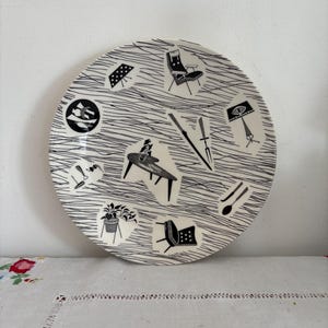 Può includere: Un piatto in ceramica bianco e nero con un design moderno di metà secolo che presenta mobili, piante e altri oggetti per la casa. Il piatto ha uno sfondo a strisce bianche e nere.