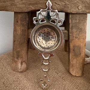 May include: A silver tea infuser with an ornate design. The infuser has a round basket with small holes and a decorative handle. The handle features a series of connected shapes. The infuser is displayed against a rustic wooden background.