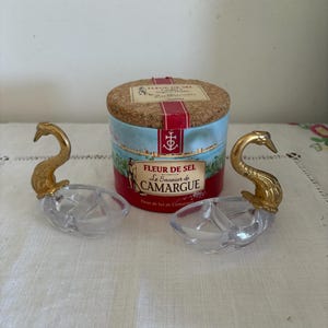 Puede incluir: Un juego de dos saleros con forma de cisne con cabezas de cisne doradas y platos de cristal transparente. Un recipiente cilíndrico de sal Fleur de Sel de Camargue está en el fondo, con tapa de corcho y etiqueta roja y azul.