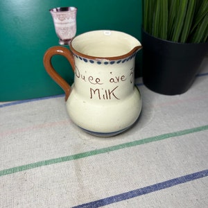 Puede incluir: Un pequeño creamer de cerámica blanca con asa marrón. Tiene ribetes azules y marrones y las palabras "Duce ave Milk" escritas en el lateral.
