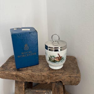 May include: A Royal Worcester egg cup with a silver lid and handle, featuring a bird and water scene. The blue box next to it has the Royal Worcester logo. The items are on a rustic wooden stool.