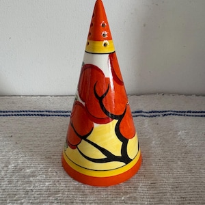May include: A ceramic cone-shaped ornament with a bright design. The cone has a red, yellow, and orange colour scheme with black outlines. The top has several small holes. The base is orange, with a yellow and black design.