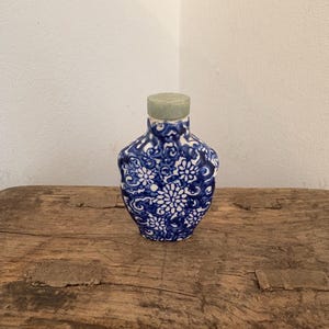 Puede incluir: Una botella de cerámica azul y blanca con un diseño floral. La botella tiene una forma pequeña y redonda con un tapón verde claro. La botella está sobre una superficie de madera rústica.