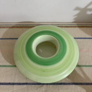 May include: A circular ceramic candle holder with bands of light and dark green. The candle holder has a central aperture and is placed on a striped fabric background.