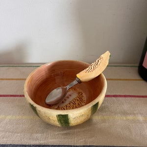 May include: A small, ceramic bowl with green stripes and a melon design. A spoon with a melon-shaped handle is inside the bowl.