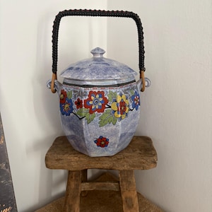 Könnte beinhalten: Ein blauer Keramik-Keksdose mit Deckel und schwarzem Griff. Die Dose ist mit bunten Blumenmustern in Rot, Blau und Gelb verziert. Sie steht auf einem kleinen, rustikalen Holzschemel.