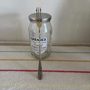 May include: A silver fork with a decorative handle stands in front of a clear glass jar with a metal lid. The jar has a label that reads "GARNER'S Original Pickled Onions". The fork has three tines and a twisted stem.