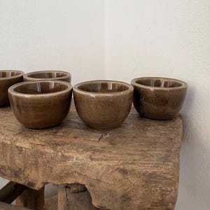 May include: A collection of small, round ceramic bowls with a glossy, dark brown glaze. Each bowl features a smooth, curved exterior and a wide, open top. The bowls are arranged in a group, showcasing their uniform shape and reflective, earthy brown finish.