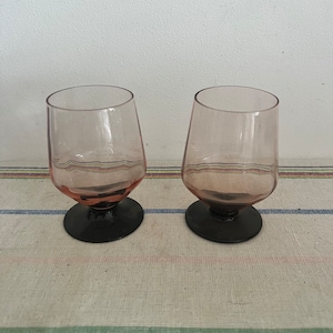 Könnte beinhalten: Zwei klare Trinkgläser aus Glas mit rosa Tönung und schwarzen Füßen. Die Gläser stehen auf einer gestreiften Tischdecke.