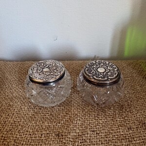 May include: Two antique glass jars with ornate silver lids. The jars have a diamond-cut pattern on the glass. The lids are decorated with detailed floral and scroll designs. The jars are displayed on a textured, brown surface.