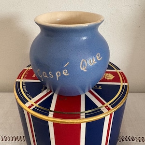 Puede incluir: Un jarrón de cerámica azul con la inscripción dorada "Gaspé Que", colocado sobre una lata redonda con un diseño de la Union Jack en rojo, blanco y azul.