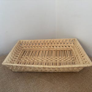 May include: A rectangular woven tray made of light beige natural fibers. The tray features a flat base with a grid-like pattern of thin vertical strips and a decorative, open-weave border along the four sides, creating a structured rim.