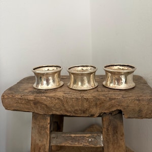 May include: Three silver napkin rings with a flared design and beaded edges are displayed on a rustic wooden stool. The rings have a polished finish, reflecting light. The stool has a weathered appearance.