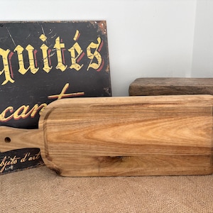 May include: A rectangular wooden serving board with a light brown natural wood grain finish and a handle featuring a circular hole. The board has rounded corners and a smooth surface, showcasing the organic texture and variations of the wood.